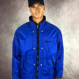 May include: A bright blue Nike jacket with a black zipper, black trim, and a black Nike logo on the chest. The jacket has a black collar and a black snap closure. The mannequin is wearing a black cap with a yellow logo.
