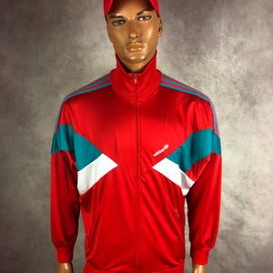 Op de afbeelding: Een rood trainingsjack met turquoise en witte accenten, met het Adidas-logo. De jas heeft een hoge kraag en een ritssluiting over de hele lengte. Er wordt ook een bijpassende rode pet gedragen.