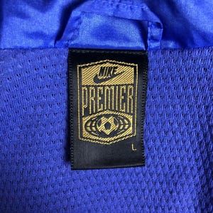 Puede incluir: Primer plano de una chaqueta deportiva azul con forro de malla. La chaqueta presenta una etiqueta Nike Premier negra y dorada con un gr&aacute;fico de bal&oacute;n de f&uacute;tbol. La etiqueta tambi&eacute;n indica la talla L.