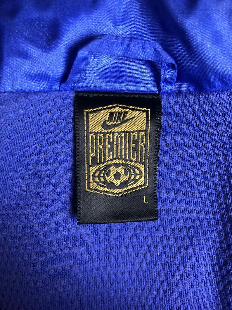 Puede incluir: Primer plano de una chaqueta deportiva azul con forro de malla. La chaqueta presenta una etiqueta Nike Premier negra y dorada con un gr&aacute;fico de bal&oacute;n de f&uacute;tbol. La etiqueta tambi&eacute;n indica la talla L.