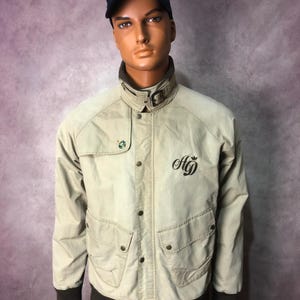 Peut inclure: Veste de bombardier beige avec col et poignets marron foncé. Elle présente une fermeture boutonnée, deux poches avant et les initiales "AG" brodées. Une casquette de baseball bleu marine avec un emblème vert et jaune est portée.