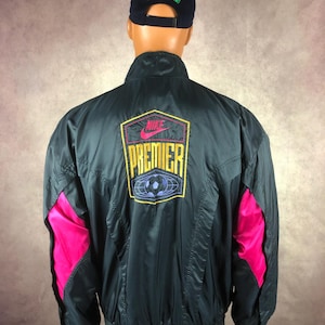 Puede incluir: Chaqueta Nike Premier verde oscuro con detalles rosas en las mangas. La chaqueta presenta el logo de Nike y el texto Premier en la espalda, junto con un gr&aacute;fico de bal&oacute;n de f&uacute;tbol. Se usa una gorra Stone Island azul marino.