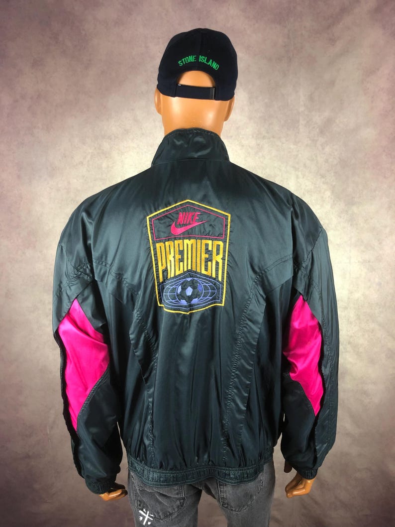 Puede incluir: Chaqueta Nike Premier verde oscuro con detalles rosas en las mangas. La chaqueta presenta el logo de Nike y el texto Premier en la espalda, junto con un gr&aacute;fico de bal&oacute;n de f&uacute;tbol. Se usa una gorra Stone Island azul marino.