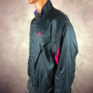 Puede incluir: Chaqueta deportiva Nike azul oscuro con logotipo rosa en el pecho y detalles rosas en las mangas. La chaqueta tiene cuello alto, cierre de cremallera y forro azul. Fabricada en material ligero, posiblemente nailon.