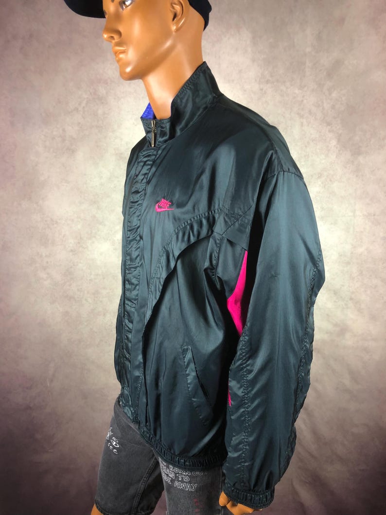 Puede incluir: Chaqueta deportiva Nike azul oscuro con logotipo rosa en el pecho y detalles rosas en las mangas. La chaqueta tiene cuello alto, cierre de cremallera y forro azul. Fabricada en material ligero, posiblemente nailon.