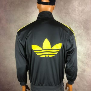 May include: Black track jacket with yellow stripes and a large yellow logo on the back. The jacket has a full zip and ribbed cuffs and hem. A black cap with green text is worn on the mannequin's head.