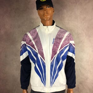 May include: A vintage Adidas track jacket in white, blue, and purple, with a full zip. The jacket features a retro design with a colour block pattern and the Adidas logo. The sleeves are navy blue. The jacket is displayed on a mannequin.