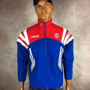 Könnte beinhalten: Eine blaue, rote und weiße Adidas-Trainingsjacke mit hohem Kragen und durchgehendem Reißverschluss. Die Jacke trägt das Adidas-Logo und das FC Bayern München-Emblem. Weiße Streifen verlaufen die Ärmel hinunter.