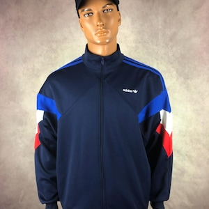 May include: Navy blue track jacket with blue and white accents on the sleeves and shoulders. The jacket has a full-length zip and the Adidas logo on the chest. A black cap with a yellow logo is worn on the mannequin's head.