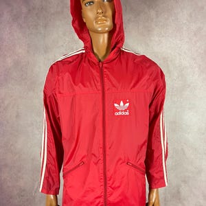 May include: A red Adidas hooded jacket with white stripes on the sleeves and shoulders. The jacket has a full-length zipper, two front pockets, and the Adidas logo on the chest. The jacket is displayed on a mannequin.