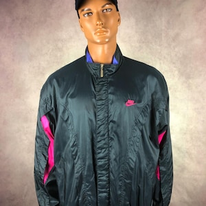 Puede incluir: Chaqueta cortavientos Nike verde oscuro con detalles rosas en las mangas y un peque&ntilde;o logotipo rosa de Nike en el pecho. La chaqueta tiene un cuello azul y una gorra de b&eacute;isbol negra.