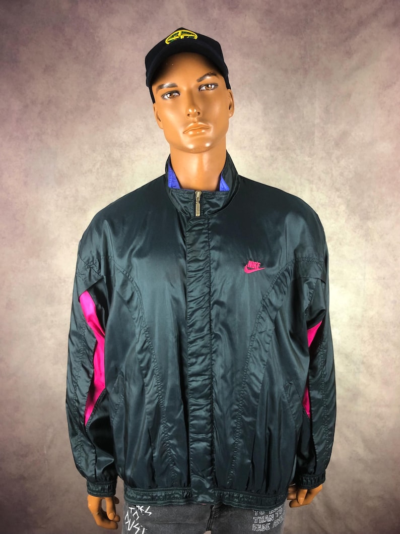 Puede incluir: Chaqueta cortavientos Nike verde oscuro con detalles rosas en las mangas y un peque&ntilde;o logotipo rosa de Nike en el pecho. La chaqueta tiene un cuello azul y una gorra de b&eacute;isbol negra.