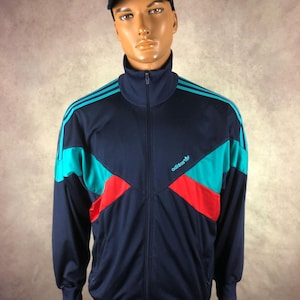 May include: Navy blue Adidas track jacket with teal and red colour block design. The jacket has a high collar and a full-length zip. The Adidas logo is visible on the chest. The mannequin is wearing a black cap with a yellow logo.