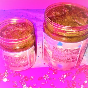 Puede incluir: Dos tarros de vidrio llenos de un exfoliante de az&uacute;car moreno. Los tarros tienen etiquetas rosas y azules con purpurina y el texto "This is all you need to get that glow". Los tarros est&aacute;n sobre una superficie rosa con gemas de pl&aacute;stico transparentes esparcidas a su alrededor.