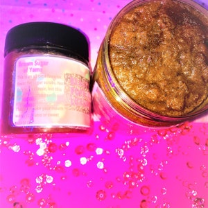 Puede incluir: Primer plano de un tarro de vidrio lleno de exfoliante de az&uacute;car moreno. El tarro tiene una etiqueta que dice "Brown Sugar Yams" y describe el producto como un sabor favorito de todos los tiempos para exfoliantes de az&uacute;car. El tarro est&aacute; sobre una superficie rosa con purpurina.