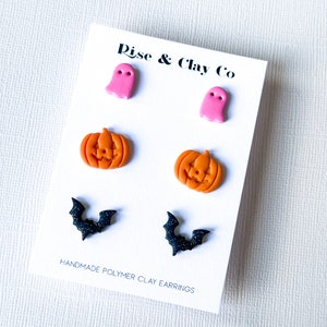 May include: A set of handmade polymer clay earrings on a white card. The earrings feature a pink ghost, orange jack-o'-lantern, and black bat with glitter. The card reads "Rise & Clay Co" and "Handmade Polymer Clay Earrings."