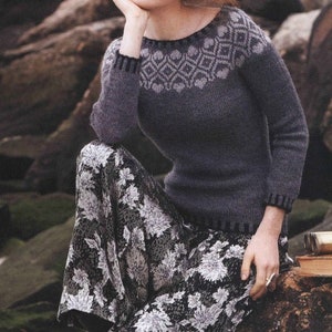 May include: A gray knitted sweater with a white geometric pattern on the neckline. The sweater has long sleeves and a relaxed fit. The model is wearing the sweater with a black and white floral skirt.