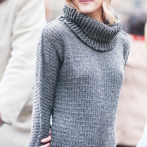 May include: A gray knitted turtleneck sweater with long sleeves. The sweater has a thick, chunky knit and a relaxed fit.