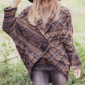 May include: A brown and gray knitted cardigan with a cable knit pattern. The cardigan has a long, loose fit and is open in the front. The cardigan is worn over a white shirt and blue jeans.