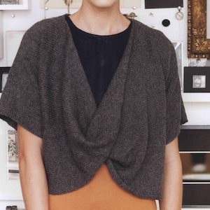 Könnte beinhalten: Ein graues Strickoberteil mit einer verdrehten Vorderseite und kurzen Ärmeln. Das Oberteil ist kurz geschnitten und hat einen V-Ausschnitt.