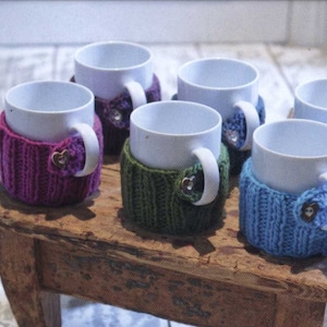 May include: Five white ceramic mugs with knitted cosy covers in different colours. The cosies are purple, green, blue, and brown. Each cosy has a heart-shaped button.