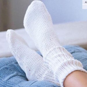 Puede incluir: Un par de calcetines blancos de punto con textura acanalada. Los calcetines están puestos y los pies descansan sobre una manta azul y blanca.