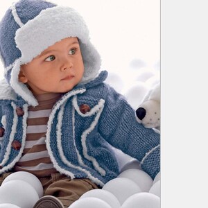 May include: A baby wearing a blue knitted jacket with white trim and a matching blue and white knitted hat. The baby is sitting in a pile of white balls.