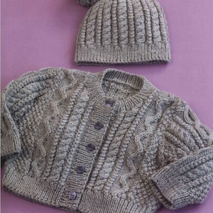 Op de afbeelding: Een grijze gebreide cardigan met een kabelbreipatroon en knopen. Het wordt gecombineerd met een grijze gebreide muts met een pompon.