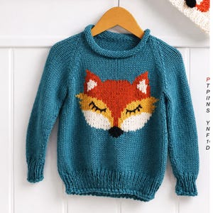 Könnte beinhalten: Ein tealblauer Strickpullover mit einem Fuchsmotiv. Der Fuchs hat orange, weiße, gelbe und schwarze Details. Der Pullover wird auf einem Holzbügel vor weißem Hintergrund präsentiert. Der Pullover hat einen gerippten Kragen und Bündchen.