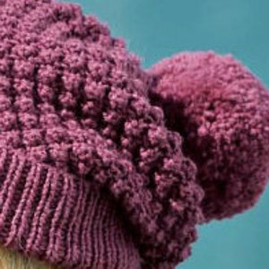 Può includere: Un berretto di lana di colore viola scuro con un grande pon pon in cima. Il berretto ha un design testurizzato e irregolare.