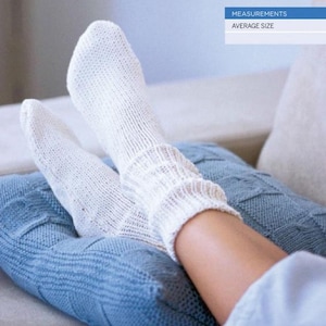 Può includere: Un paio di calzini bianchi lavorati a maglia con polsini a coste. I calzini vengono indossati mentre ci si rilassa su una coperta blu lavorata a maglia. L'immagine mostra i calzini e le gambe di una persona.