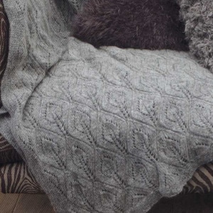 Könnte beinhalten: Eine graue gestrickte Wolldecke mit einem Rautenmuster und einem zarten Blattdesign. Die Decke liegt über einem braunen und weißen gestreiften Sofa.