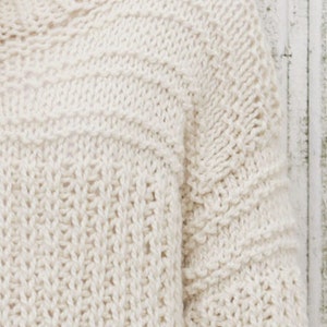 May include: A close-up of a cream-colored knitted sweater with a textured stitch pattern.