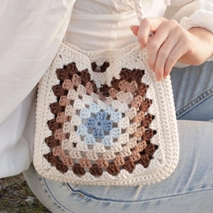 May include: A cream-coloured, crocheted shoulder bag with a square granny square design. The centre of the bag features a blue floral pattern, surrounded by concentric squares of brown, tan, and cream. The bag has a long, cream-coloured strap.