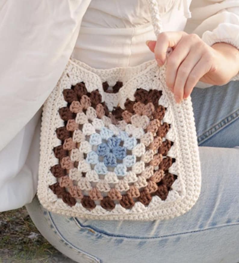 May include: A cream-colored, crocheted shoulder bag with a square granny square design. The center of the bag features a blue floral pattern, surrounded by concentric squares of brown, tan, and cream. The bag has a long, cream-colored strap.