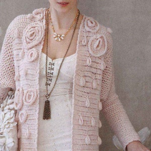 May include: A pink crocheted cardigan with a delicate floral design. The cardigan features a tassel necklace and is open at the front.