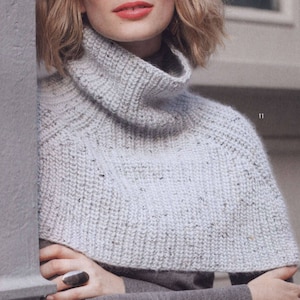May include: A light gray knitted cowl neck sweater with a textured pattern. The sweater is worn by a person with short blonde hair.