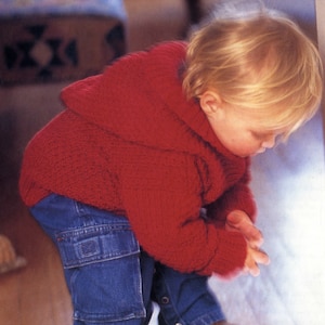 Puede incluir: Un niño pequeño con un suéter rojo de punto con capucha y pantalones vaqueros azules. El niño está mirando hacia abajo a algo fuera del encuadre.