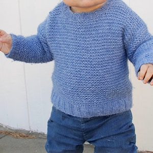 Könnte beinhalten: Ein hellblauer Strickpullover für ein Baby oder Kleinkind. Der Pullover hat einen Rundhalsausschnitt und lange Ärmel.
