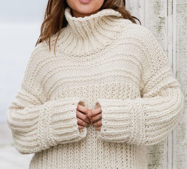 May include: A cream-colored knitted sweater with a high turtleneck and long sleeves. The sweater has a chunky knit texture and a relaxed fit.