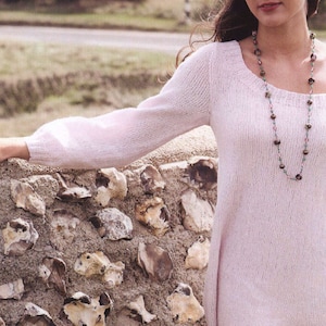 Peut inclure: Une femme portant un pull rose à épaules dénudées avec un collier de perles. Elle est debout devant un mur en pierre.