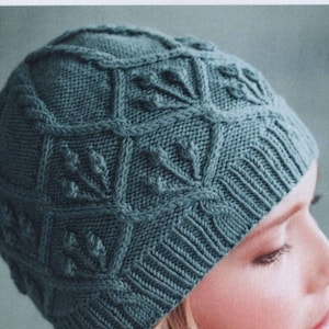 Könnte beinhalten: Eine teal-farbene Strickmütze mit einem strukturierten, geometrischen Muster. Die Mütze hat einen gerippten Saum und ein detailliertes Design aus Quadraten und blattartigen Formen. Die Mütze ist aus weichem, warmem Material gefertigt.