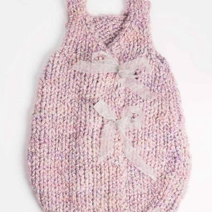 Puede incluir: Un saco de dormir para bebé de punto rosa con una cinta de satén blanca atada alrededor del cuello.