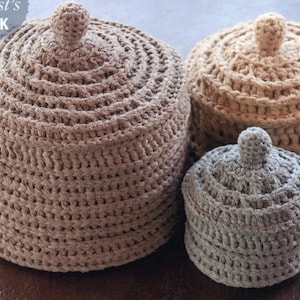 May include: Three crocheted storage containers with lids. The largest container is brown, the medium container is tan, and the smallest container is gray.