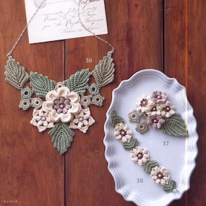May include: A crocheted jewelry set featuring a necklace, bracelet, and brooch. The necklace has a floral design with green leaves and a large central flower. The bracelet and brooch also have floral patterns. The set is made of cream, green, and purple yarn.