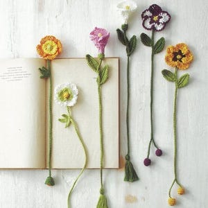 May include: A collection of handmade crochet flower bookmarks in various colors, including yellow, orange, pink, white, and purple. Each bookmark features a detailed flower design with green leaves and a long, green stem. Some have tassels or small berry accents.