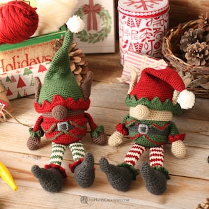 May include: Two crocheted elf ornaments with green hats, red and white striped legs, and brown faces. The elves are sitting on a wooden surface with a variety of Christmas decorations.