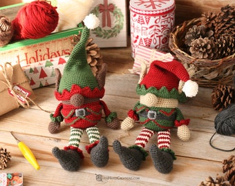 May include: Two crocheted elf ornaments with green hats, red and white striped legs, and brown faces. The elves are sitting on a wooden surface with a variety of Christmas decorations.