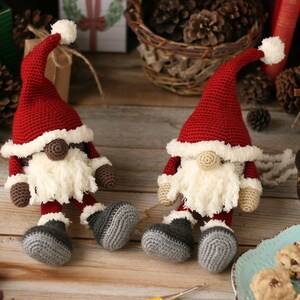 A brown and a white Crochet Santa Gnome sitting side by side surrounded by pinecones, packages, a plate of cookies, a glass of milk, and other Christmas paraphernalia.