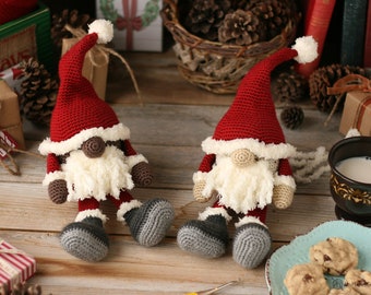 May include: Two crocheted gnomes with red hats, white beards, and gray boots. The gnomes are sitting on a wooden surface with a cup of milk and cookies in the background.